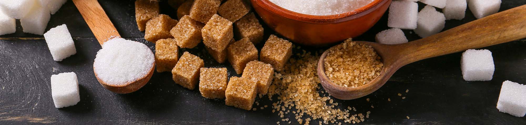 A bowl of white sugar and a pile of brown sugar cubes arranged on a dark surface.